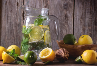 Glass pitcher of homemade lemonade, served with whole and sliced lemons, limes and mint over wooden table with wooden background.